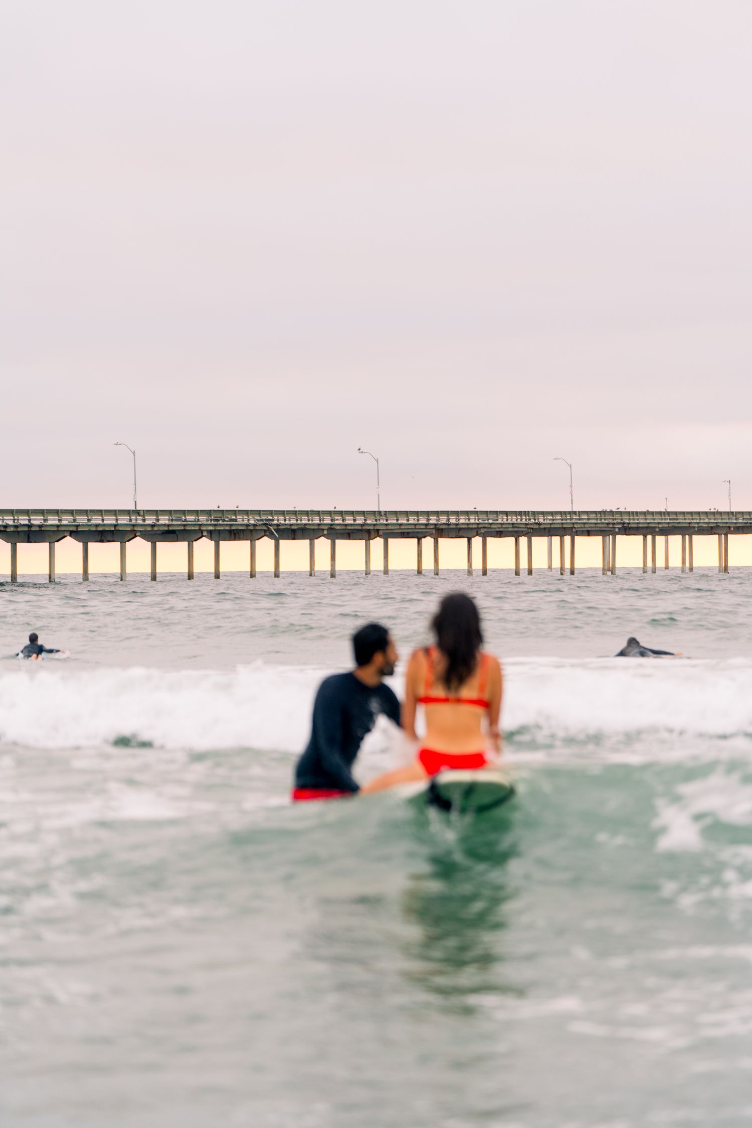 Surfers sitting in ocean water watch waves near a long coastal pier during sunset with soft pink sky.