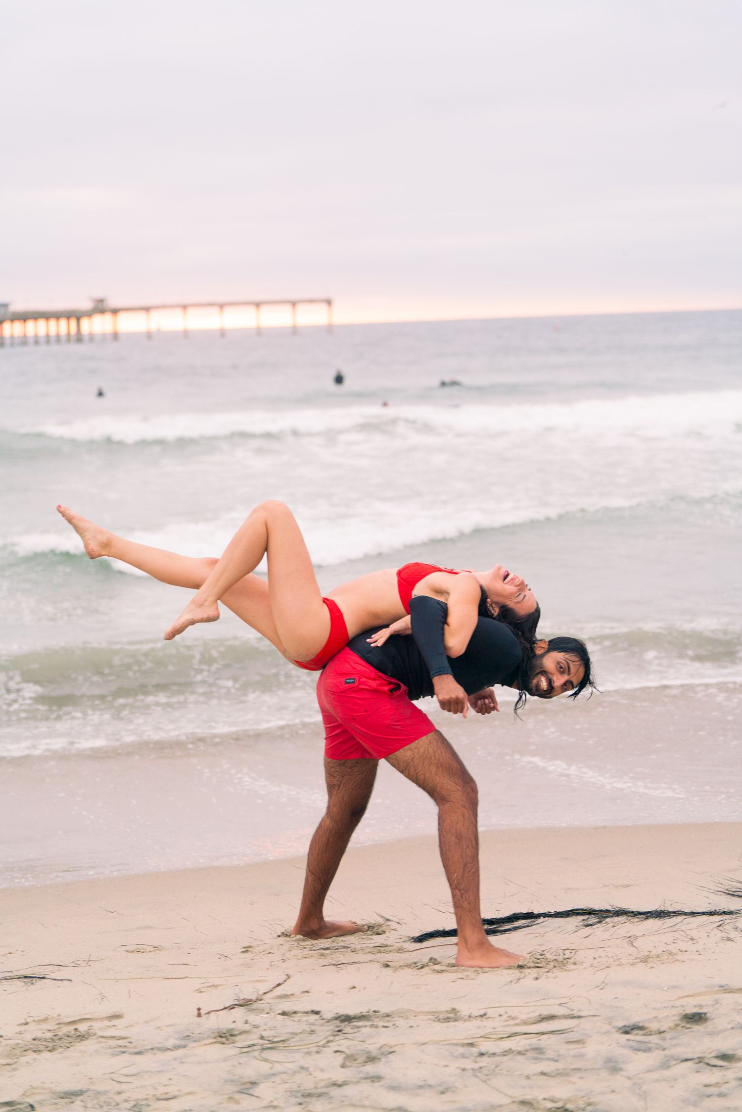 Couple performs acrobatic dance pose on beach with pier visible in background during sunset.
