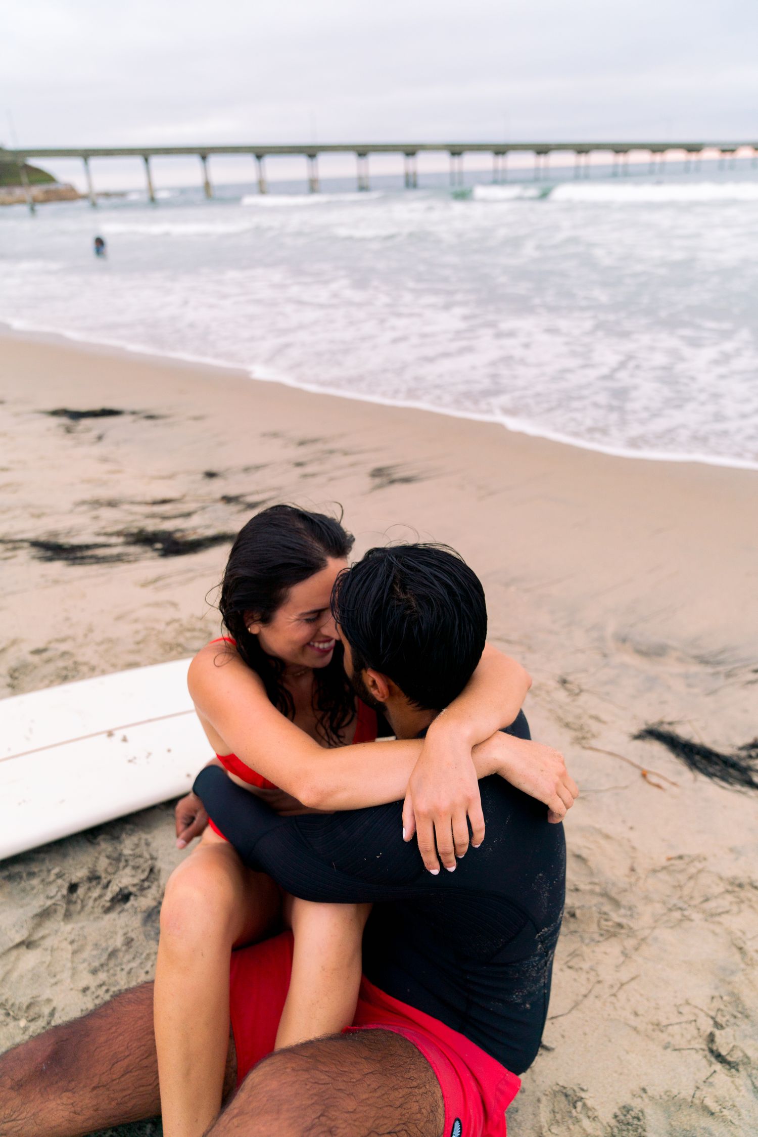 A couple embraces on a sandy beach with a long pier visible in the background on a cloudy day.