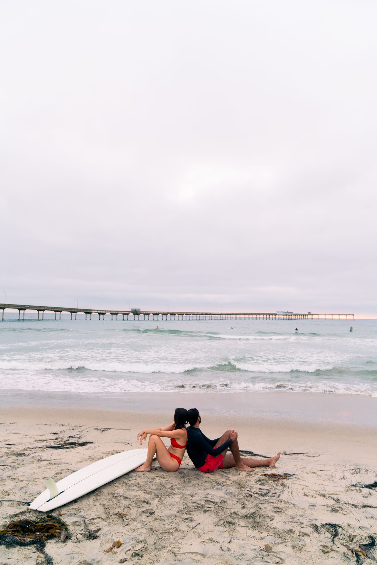 Couple relaxing on sandy beach with surfboard beside long coastal pier at dusk.