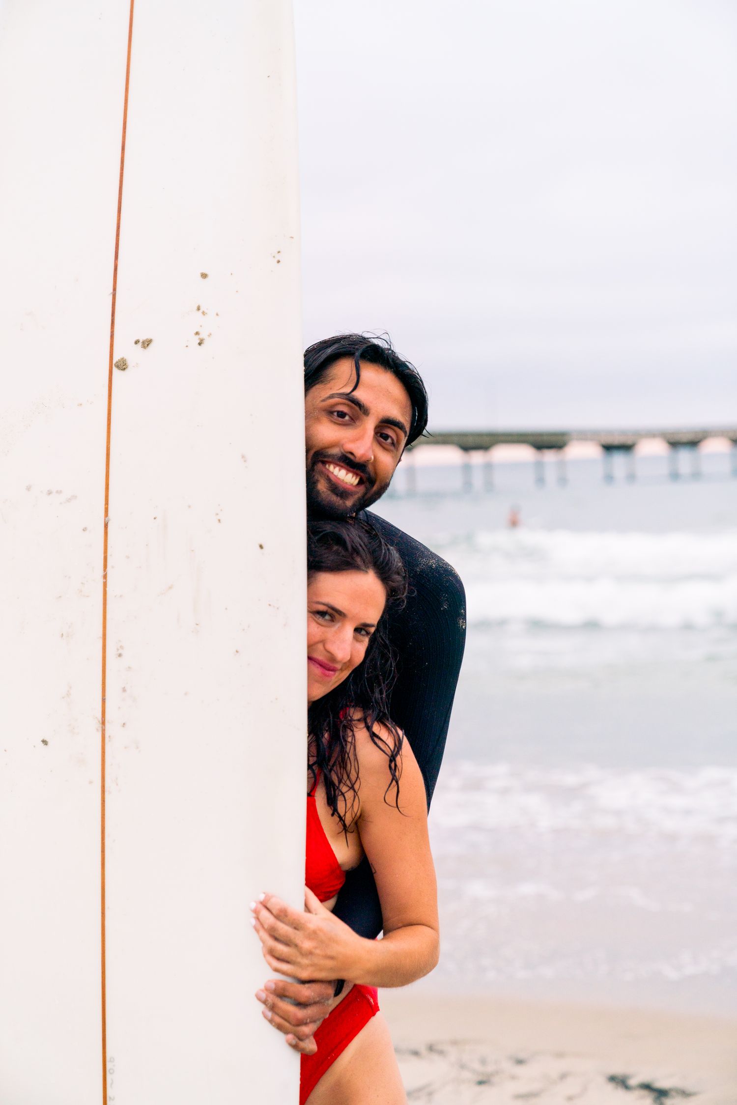 A couple peeks out from behind a white surfboard at the beach with a pier in the distance.