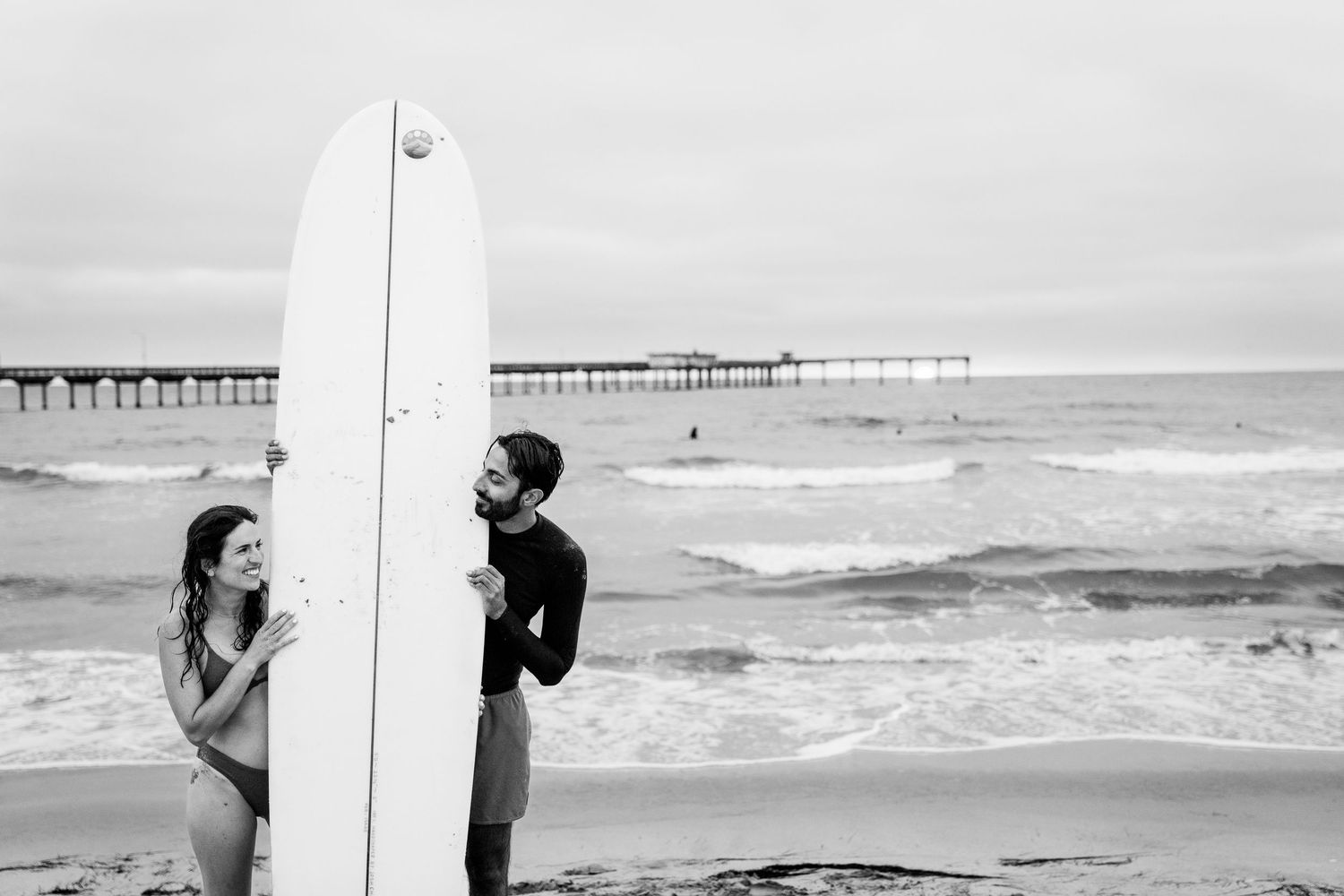 Two people stand on a beach near a surfboard with a pier visible in the background in black and white.