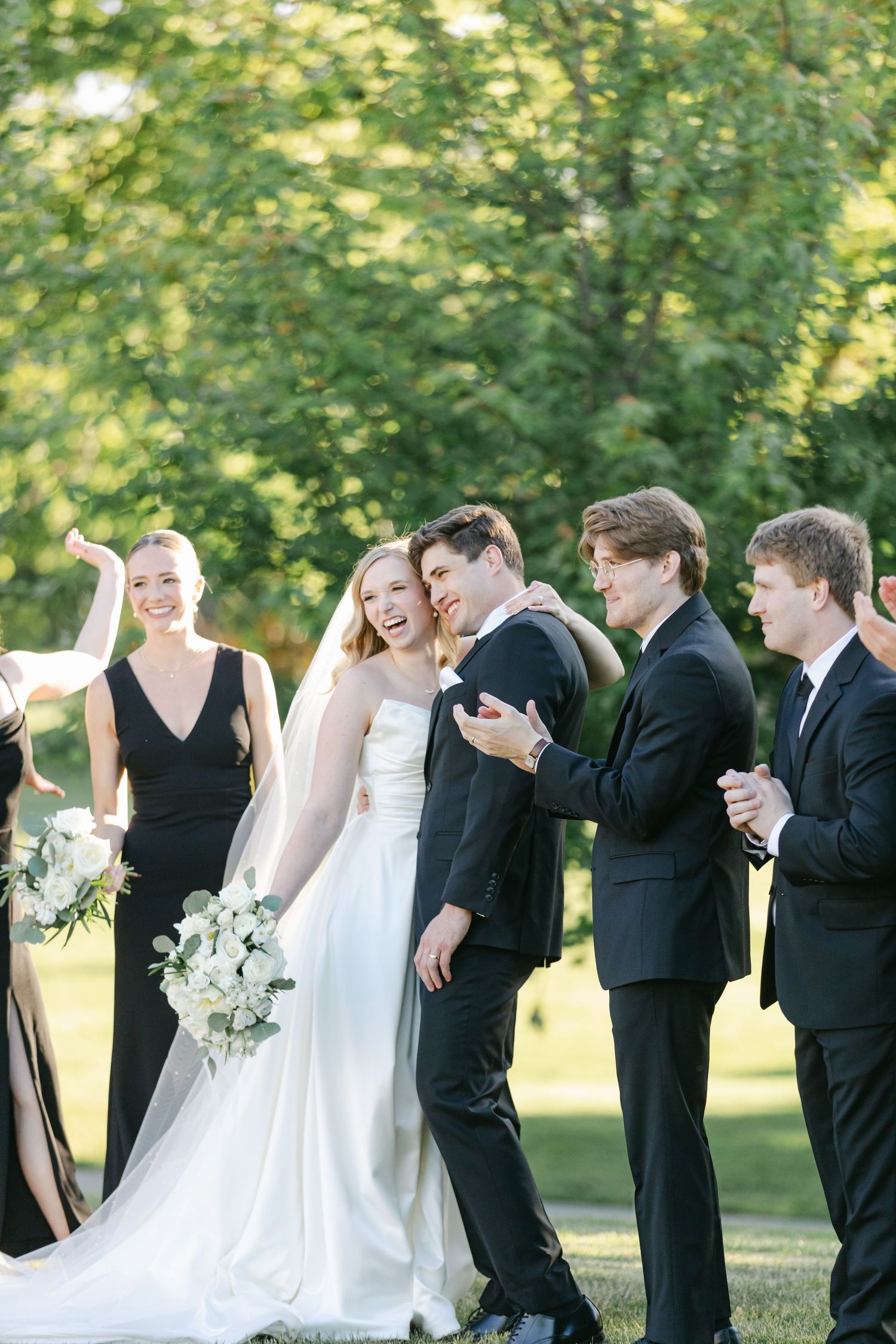 Wedding party shares a candid joyful moment together outdoors with bridesmaids in black and groomsmen in tuxedos.