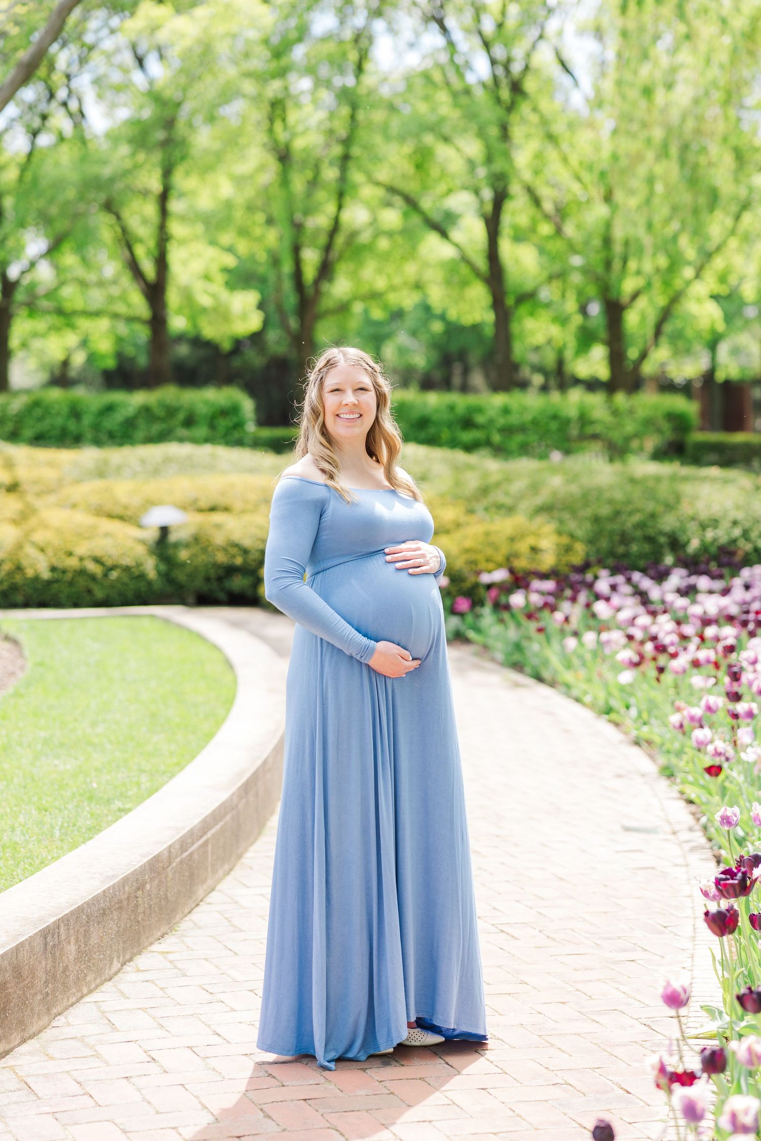 An expectant mother in a flowing light blue maxi dress poses along a garden path with blooming pink tulips at Chicago Botanic Garden.