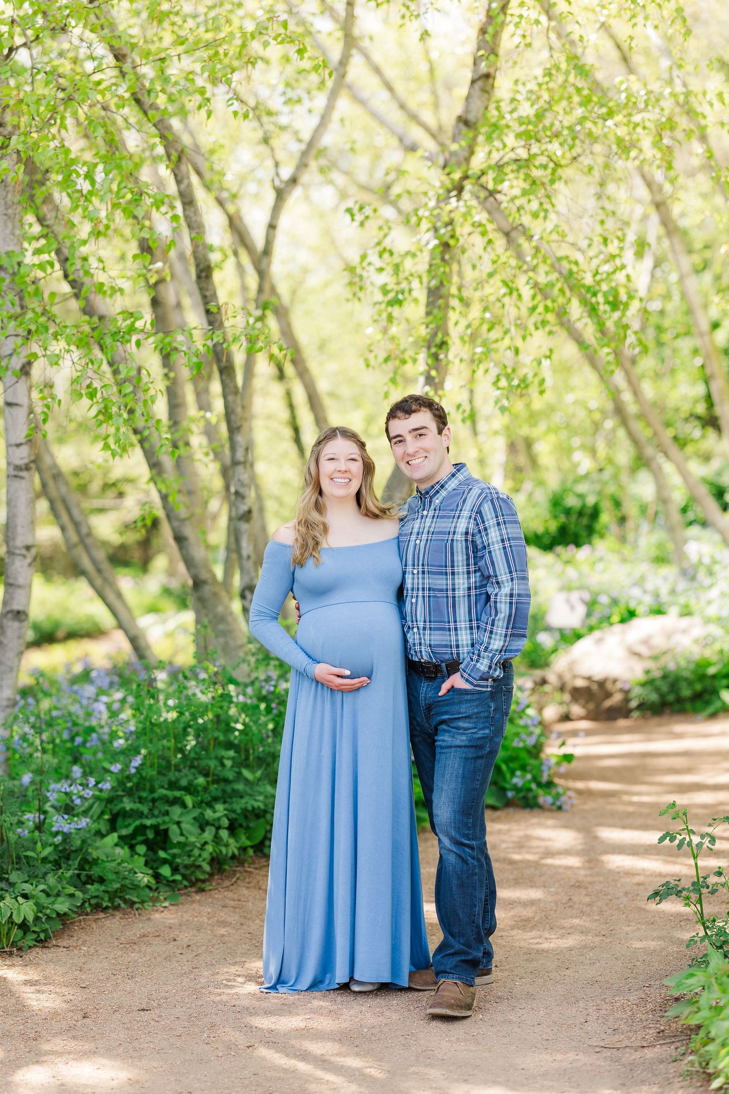 A soft blue maternity dress glows in a sunlit forest path surrounded by spring blooms and birch trees at Chicago Botanic Garden