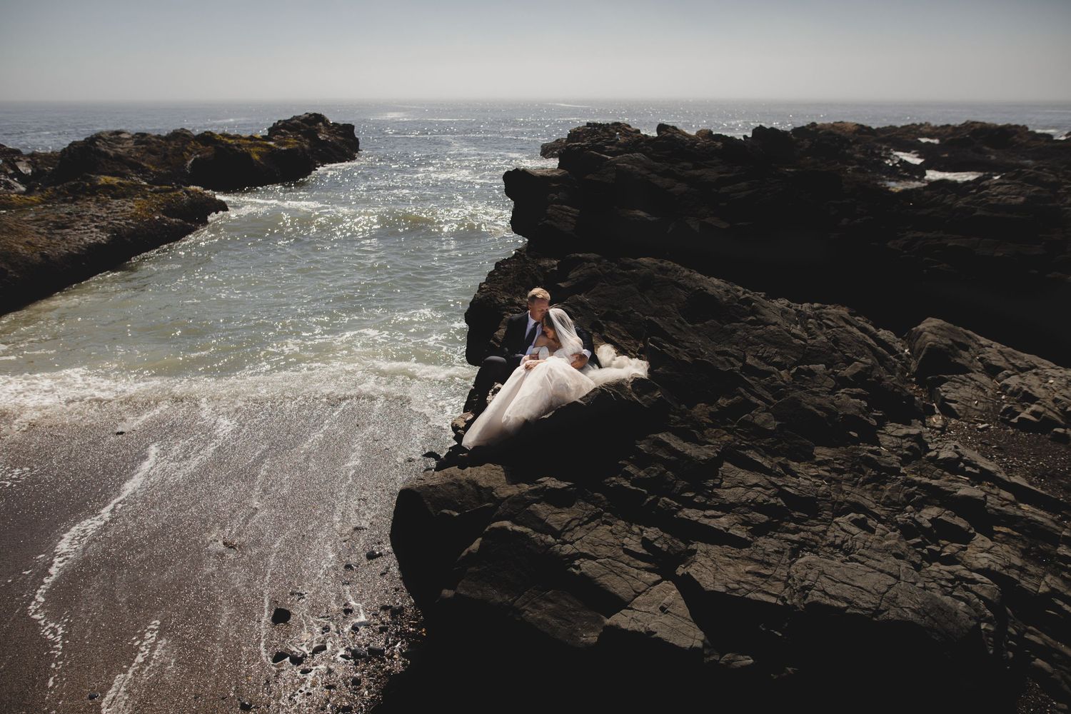 Two figures in wedding attire embrace on dramatic rocky coastline as ocean waves crash against black volcanic rocks.