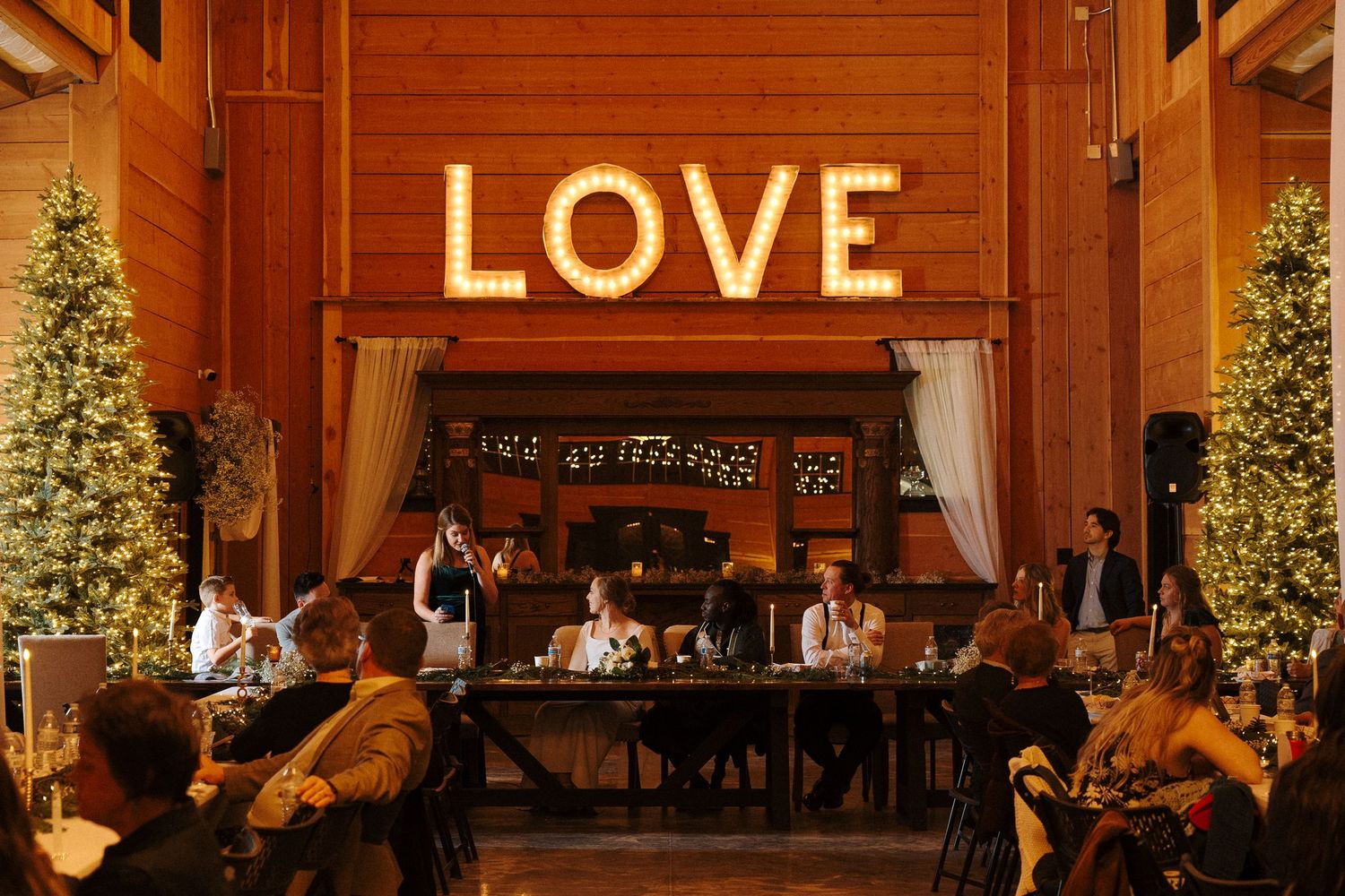 A rustic wedding reception venue with illuminated LOVE sign, Christmas trees, and guests seated at long tables.