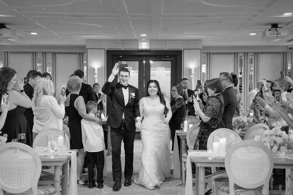Wedding guests gather at an indoor reception venue with white decor and table settings.