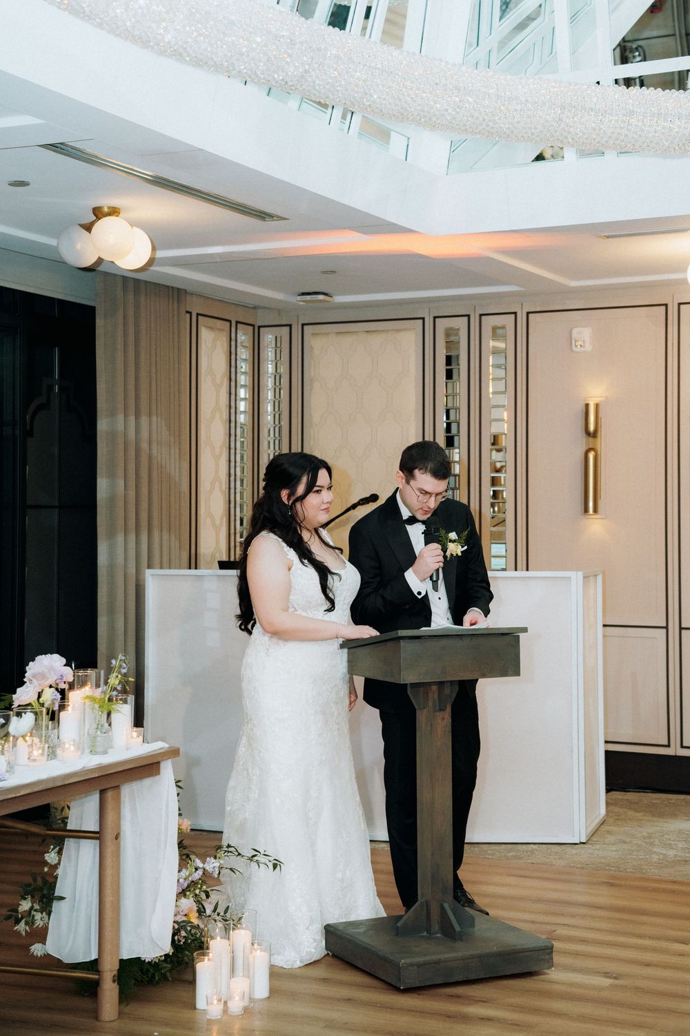 A couple at their wedding ceremony reading vows from a rustic wooden podium with candles and floral decor.