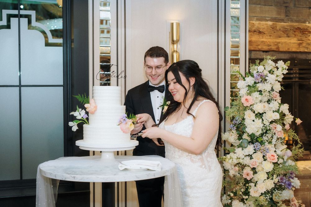 Newlyweds sharing moment while cutting their white tiered wedding cake beside floral arrangements.