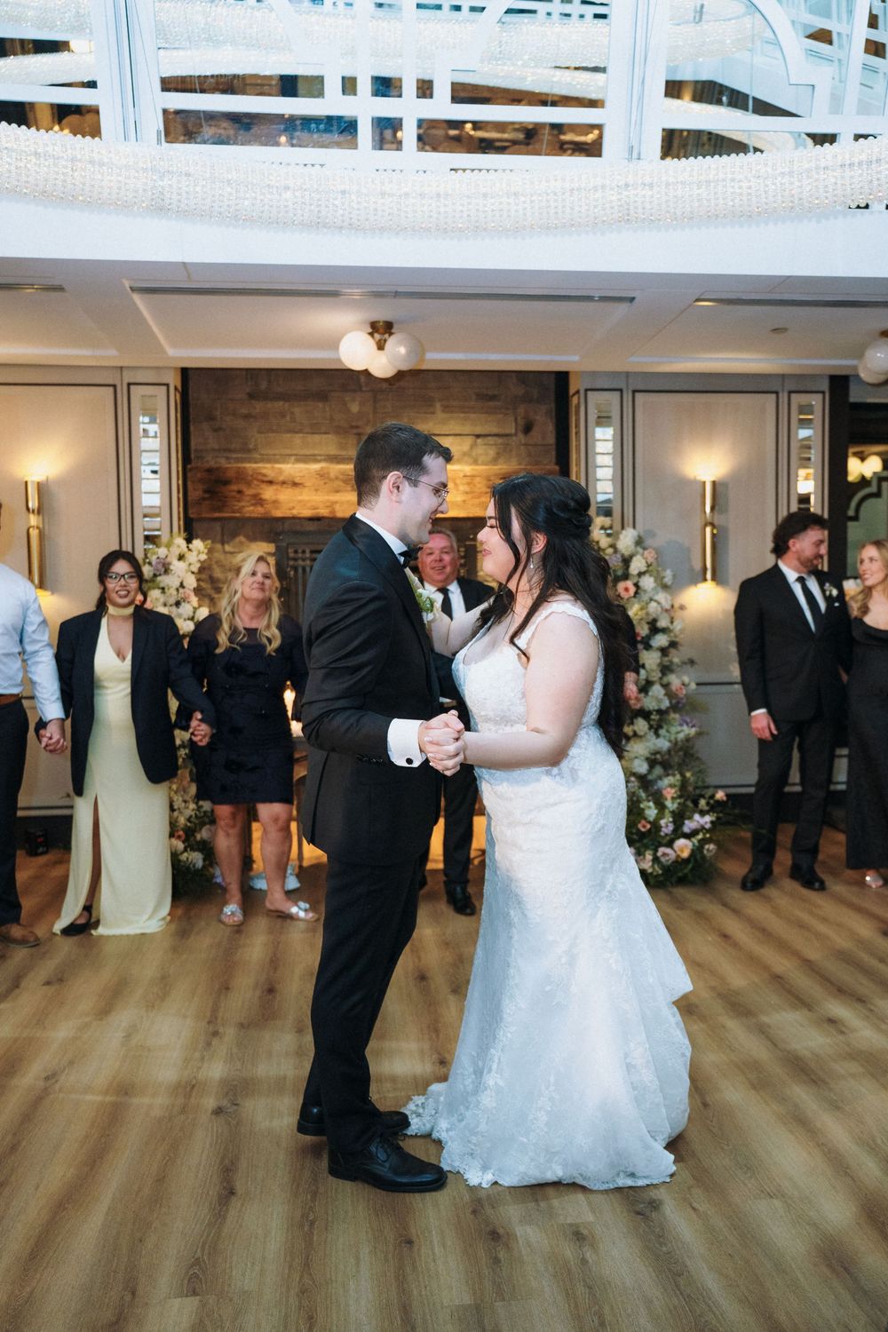First dance moment between couple on wooden dance floor with floral arch and guests watching.