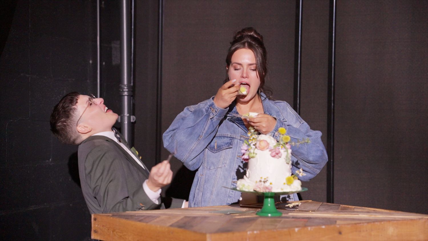 Two people sit at a wooden table laughing and eating cake together in a dimly lit space.