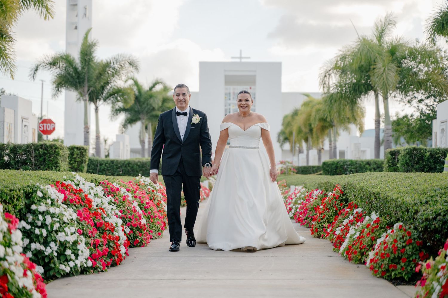 Wedding couple walks down palm-lined garden path with red and white flowers blooming alongside the walkway.