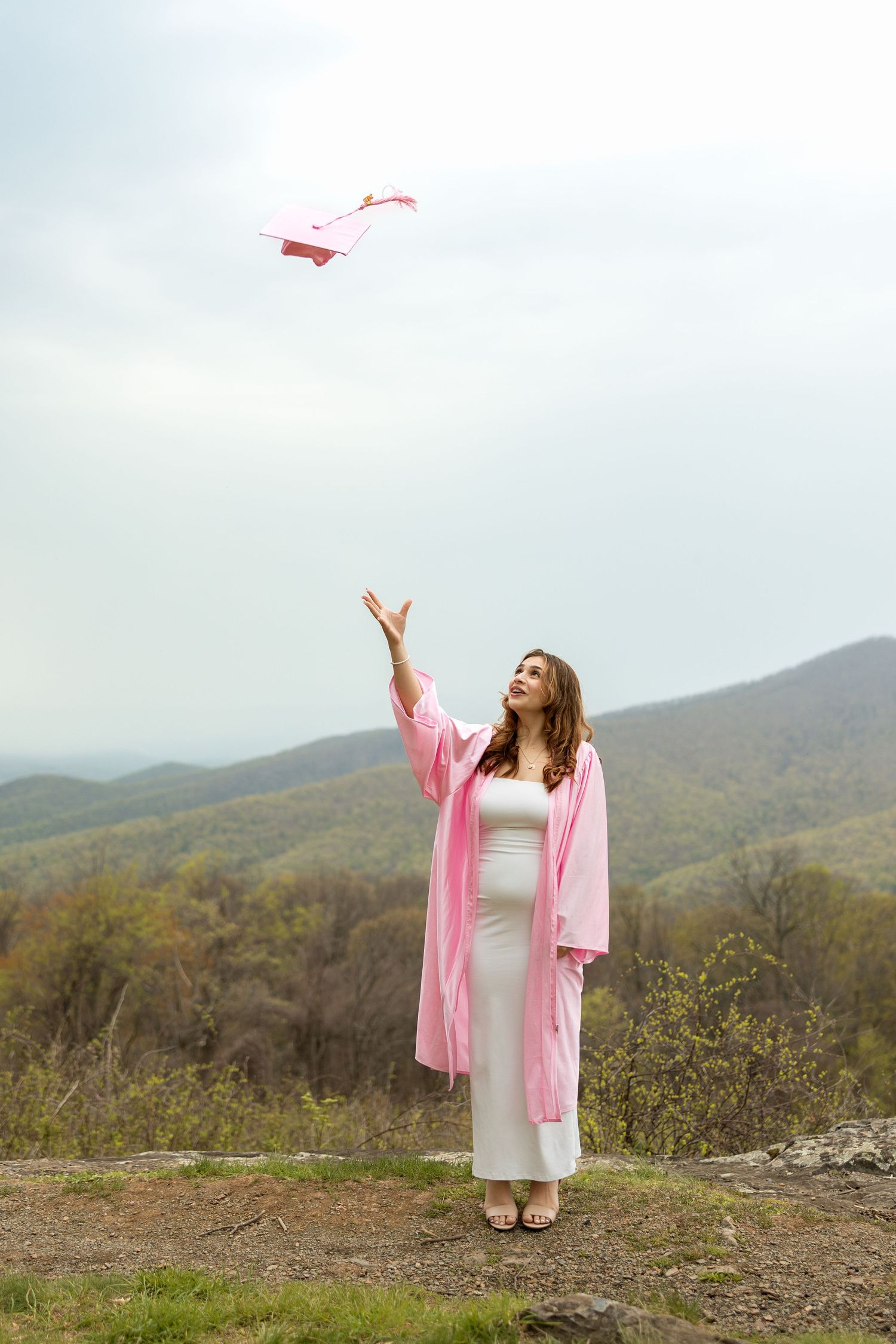 Someone in a pink robe and white dress tosses paper airplanes against a scenic mountain backdrop on a cloudy day.