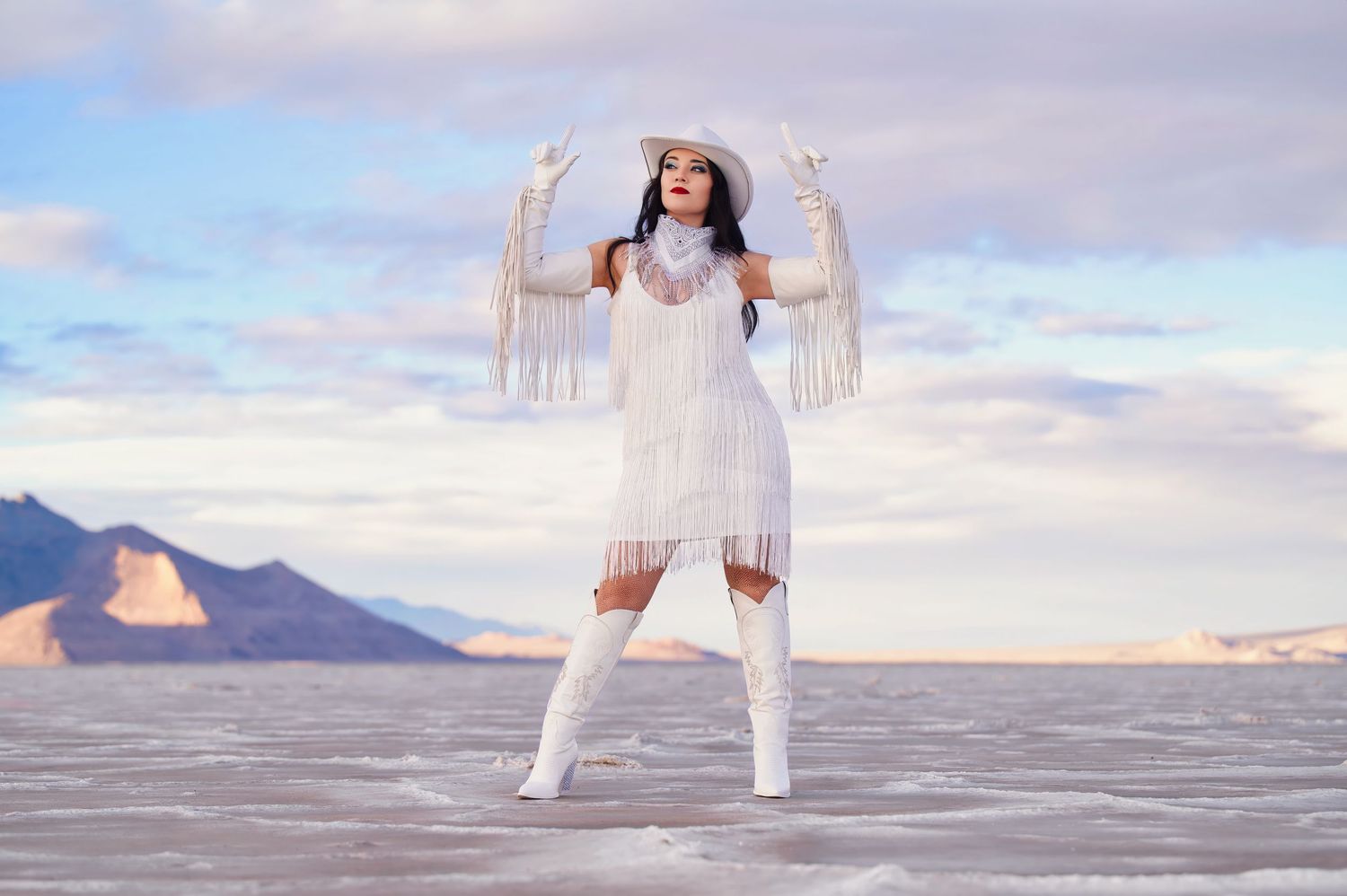 Woman in a white fringe dress and cowboy hat strikes a confident pose at sunset on salt flats. Photo by Carolina Waldroff Photography.