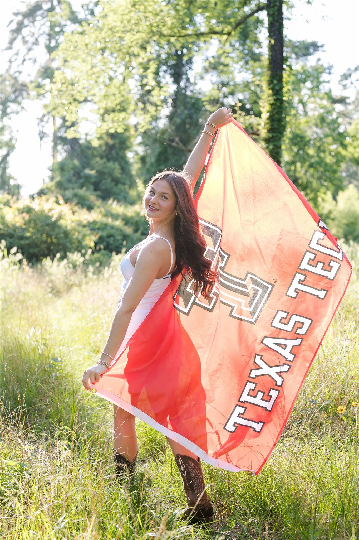 Someone holds a Texas Tech University flag while standing in a sunny meadow with trees in the background.