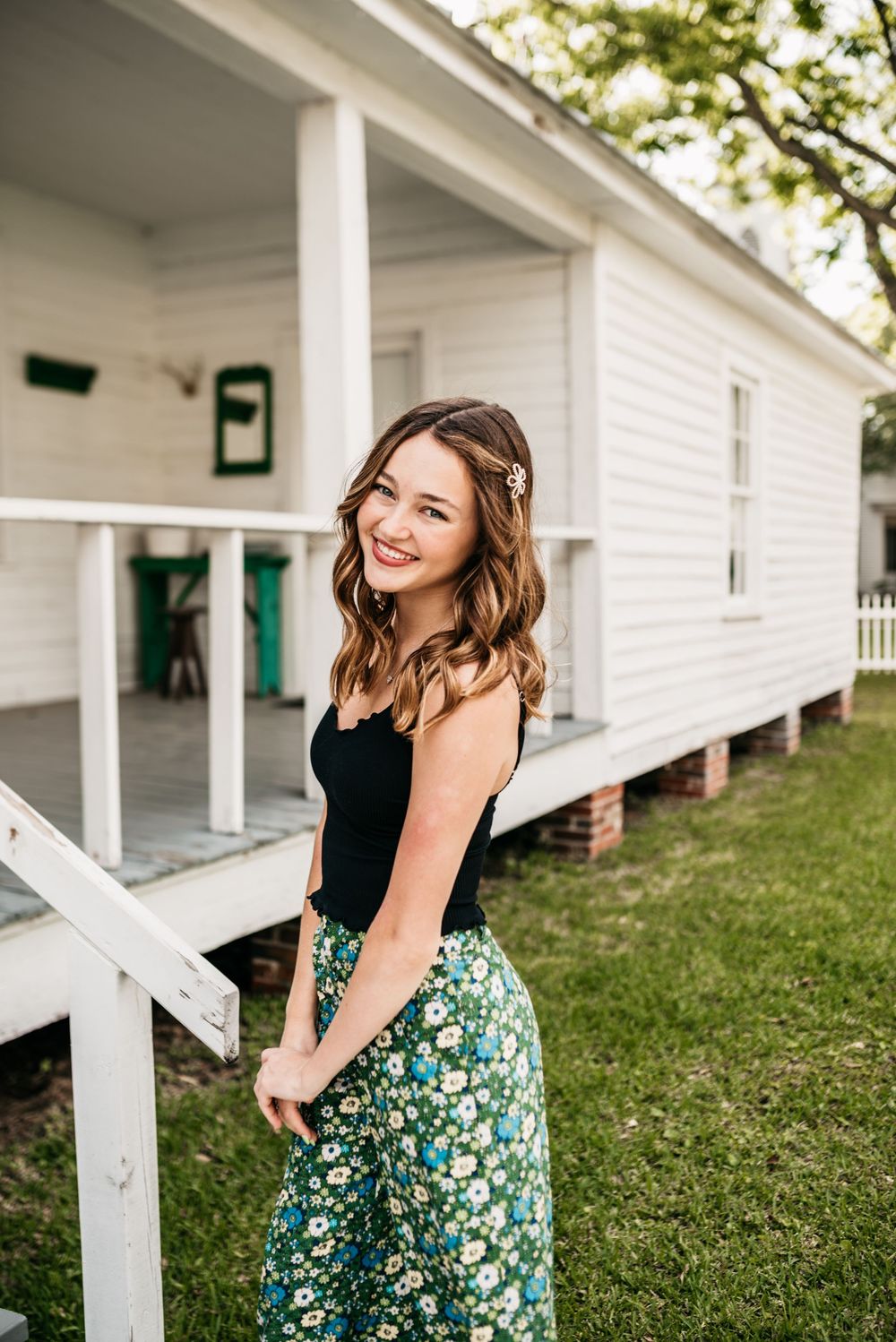 A person in a black top and floral skirt stands smiling beside a white porch railing on a summer day.