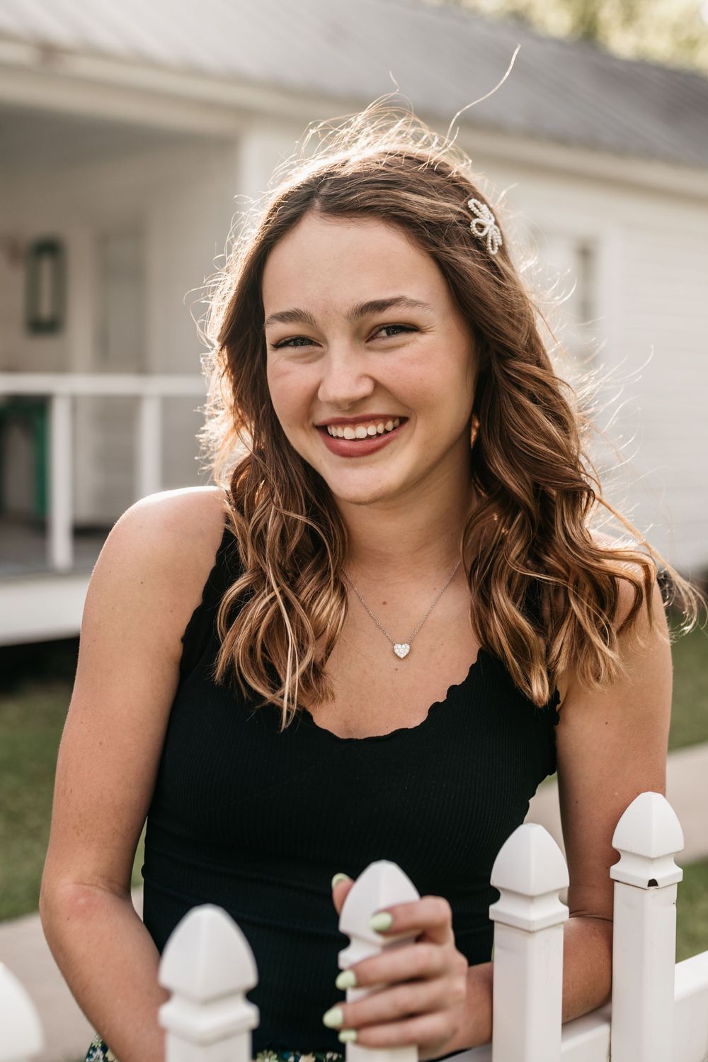 A joyful woman wearing a black tank top and delicate tiara stands by a white picket fence on a sunny day.
