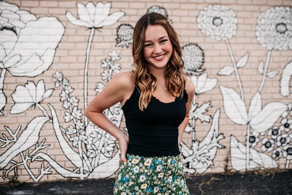 A model in a black tank top and floral skirt poses in front of a wall with painted white flower murals.