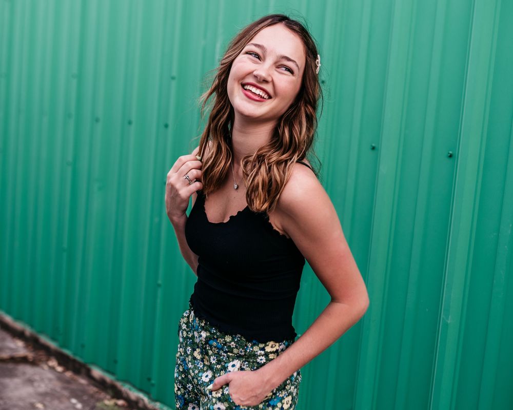 A person in a black top and floral skirt stands joyfully against a bright green corrugated metal wall.