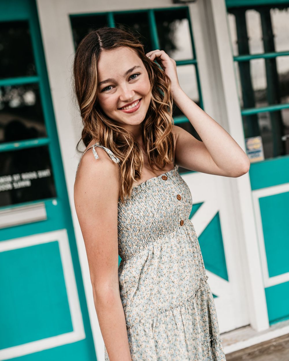 A model in a floral dress poses against a turquoise storefront with a bright, natural smile.