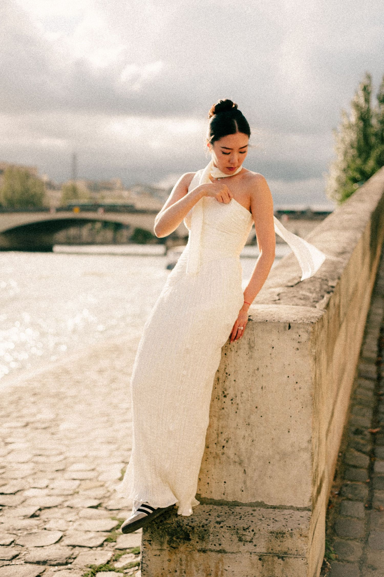 A bride in white dress poses on stone wall by river under cloudy sky.