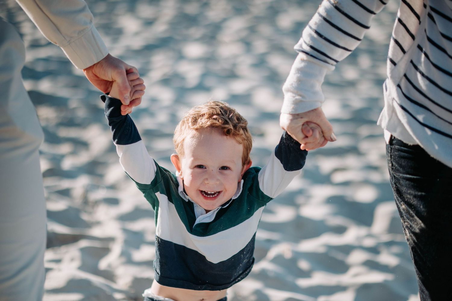 Young boy in white and navy shirt smiles while holding hands with adults on sandy beach.