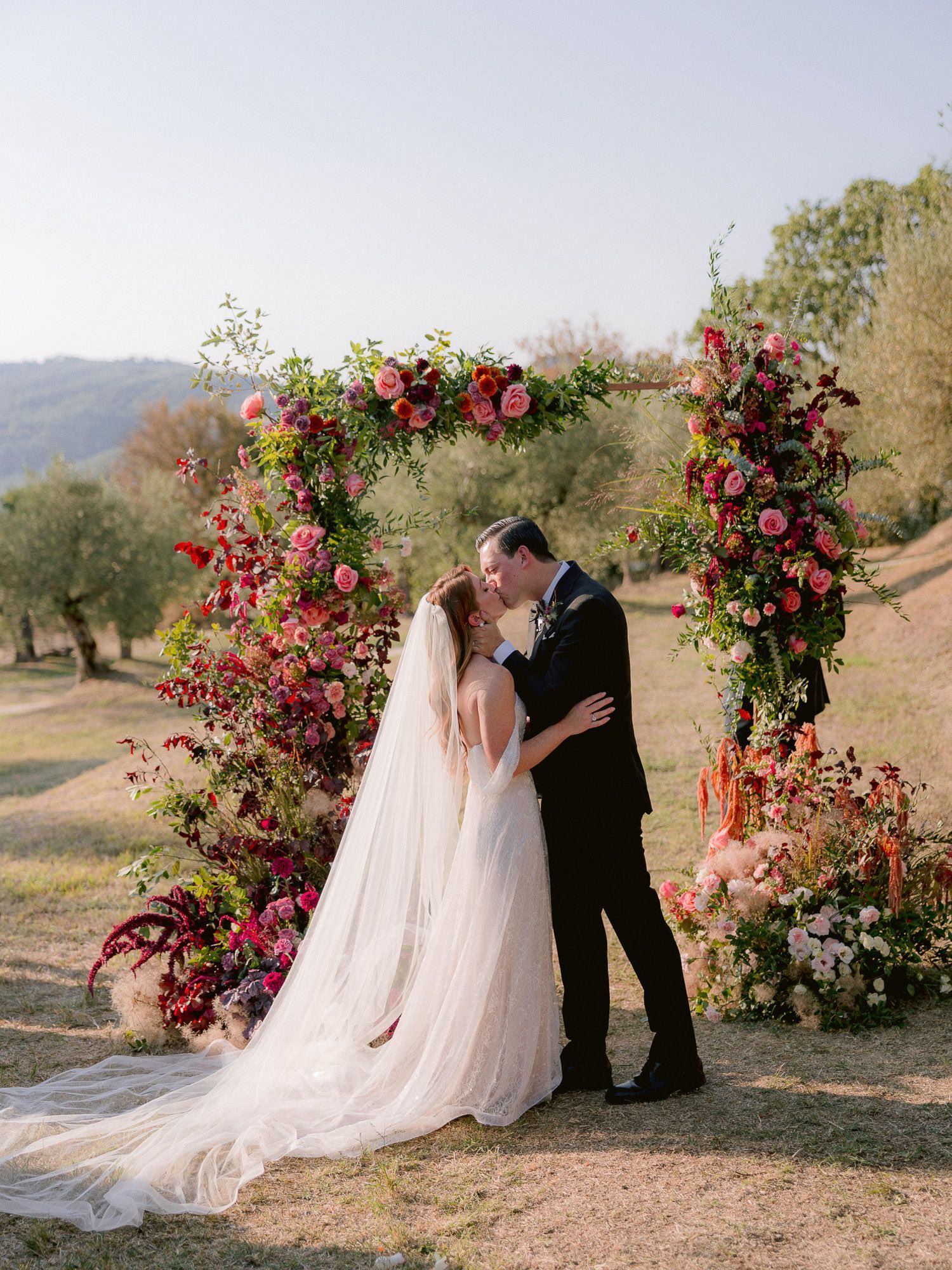 A romantic wedding couple share a kiss under an autumn-inspired floral arch with red and pink blooms in a rustic outdoor setting.
