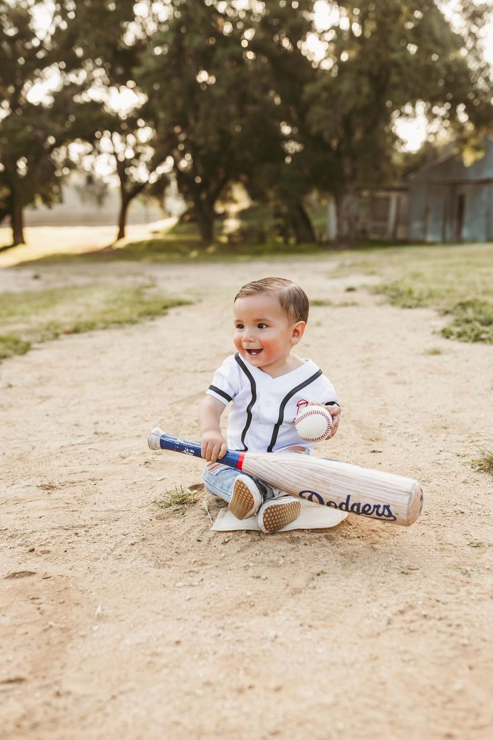 A young child wearing white baseball attire sprawls playfully on the ground during a sports photo session at sunset.