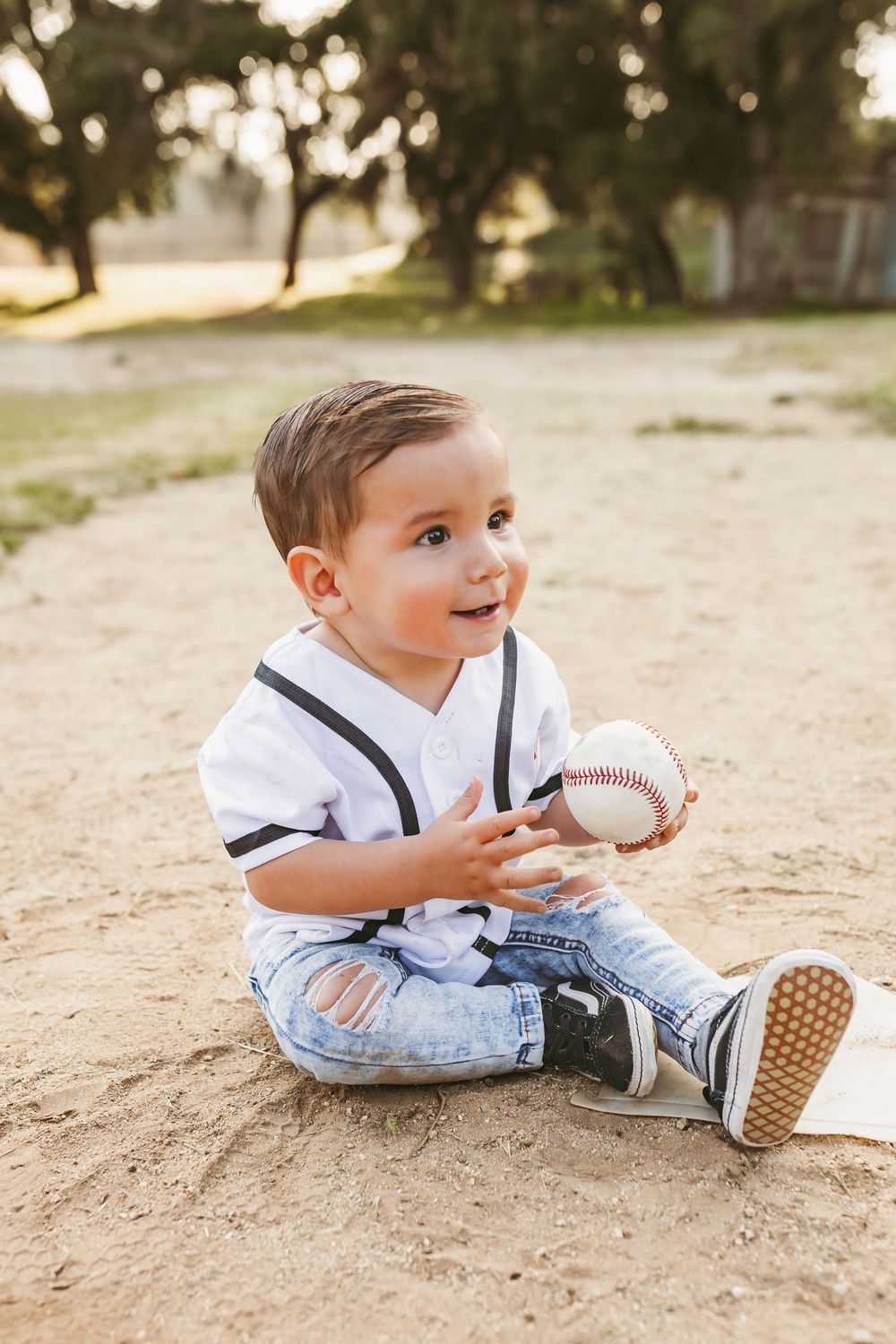 A toddler in overalls plays with a baseball on a sandy ground in warm sunlight.