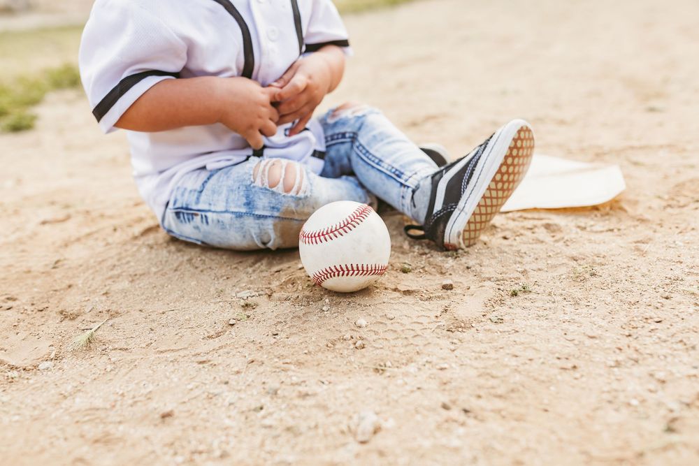 A baseball rests on dirt ground next to small legs wearing jeans and sneakers in a youth sports setting.