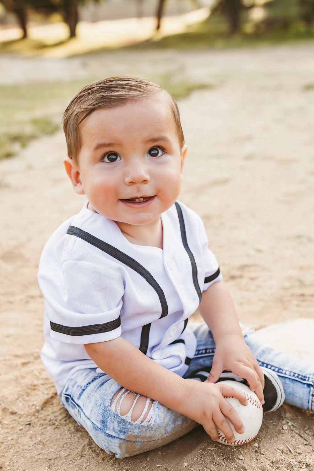 Toddler in white shirt and distressed jeans sits on the ground holding a baseball on a sunny day.