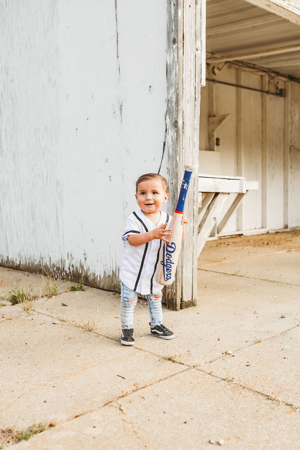 Small child in white t-shirt and stripes stands outside holding baseball bat near white building wall.