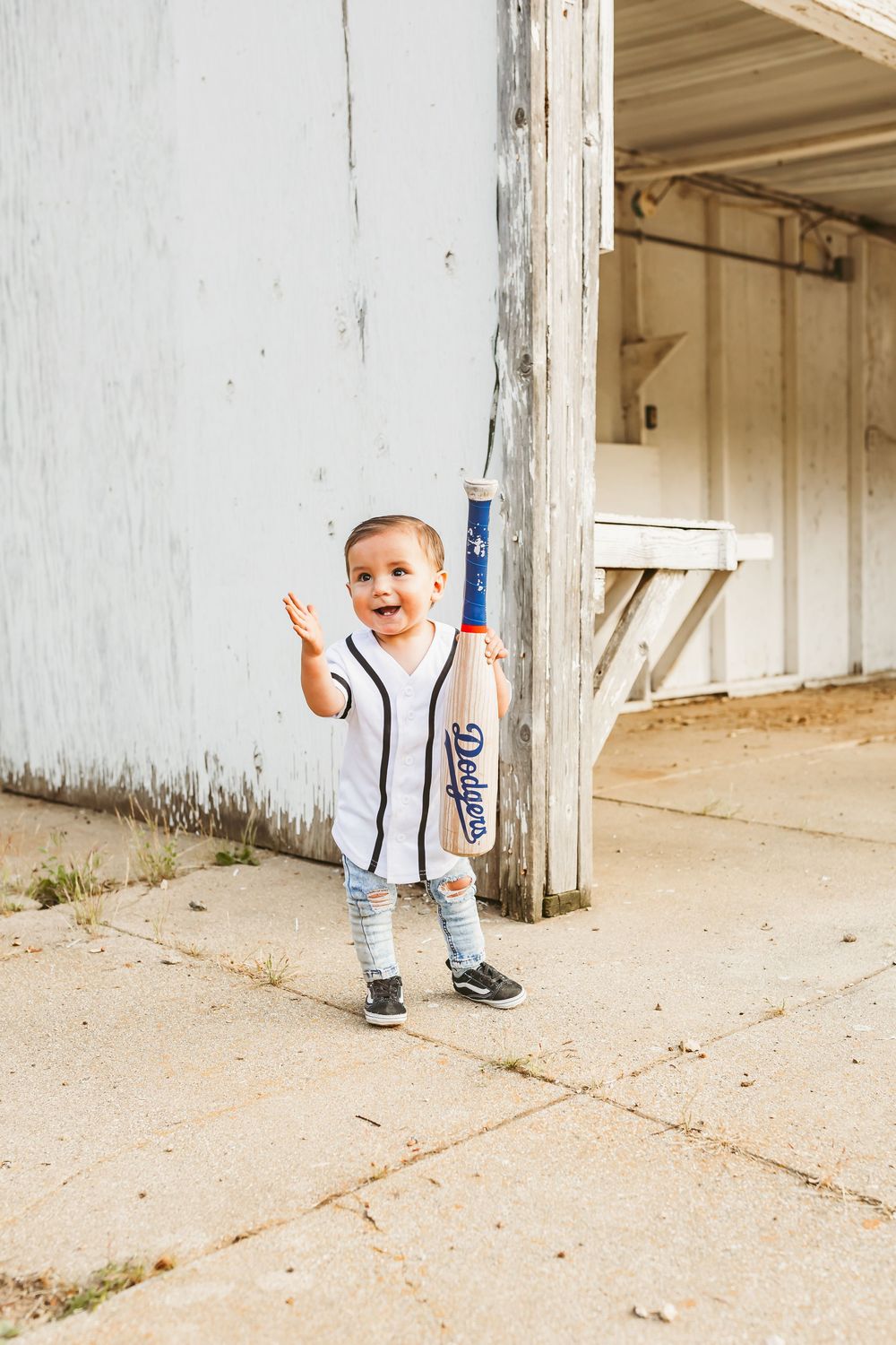 A small child in a white baseball jersey and striped socks stands playfully by a concrete pillar outside a building.