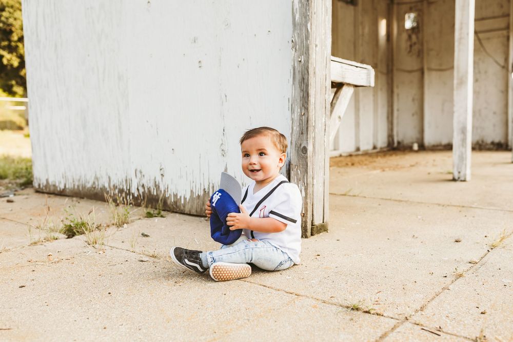 A small toddler in a blue and white outfit sits on concrete and smiles in front of a white building.