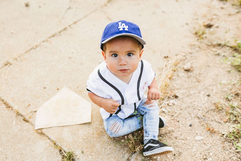 Small baseball fan in LA Dodgers cap and white jersey sits near home plate base on dirt path.