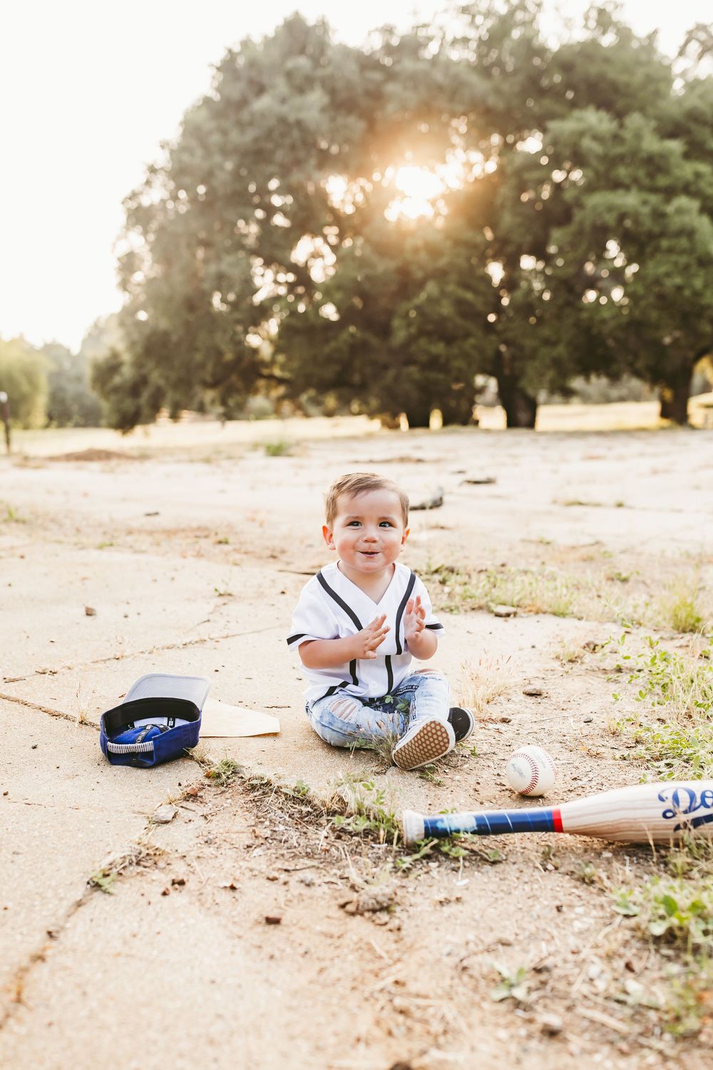 Baseball player laying in dirt field looking happy with baseball bat and gloves against sunny summer backdrop.