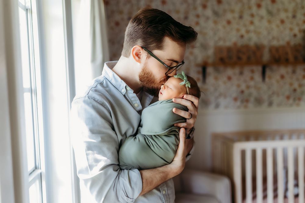A tender moment shared between parent and baby in soft natural window light, wearing gray and sage green clothing.