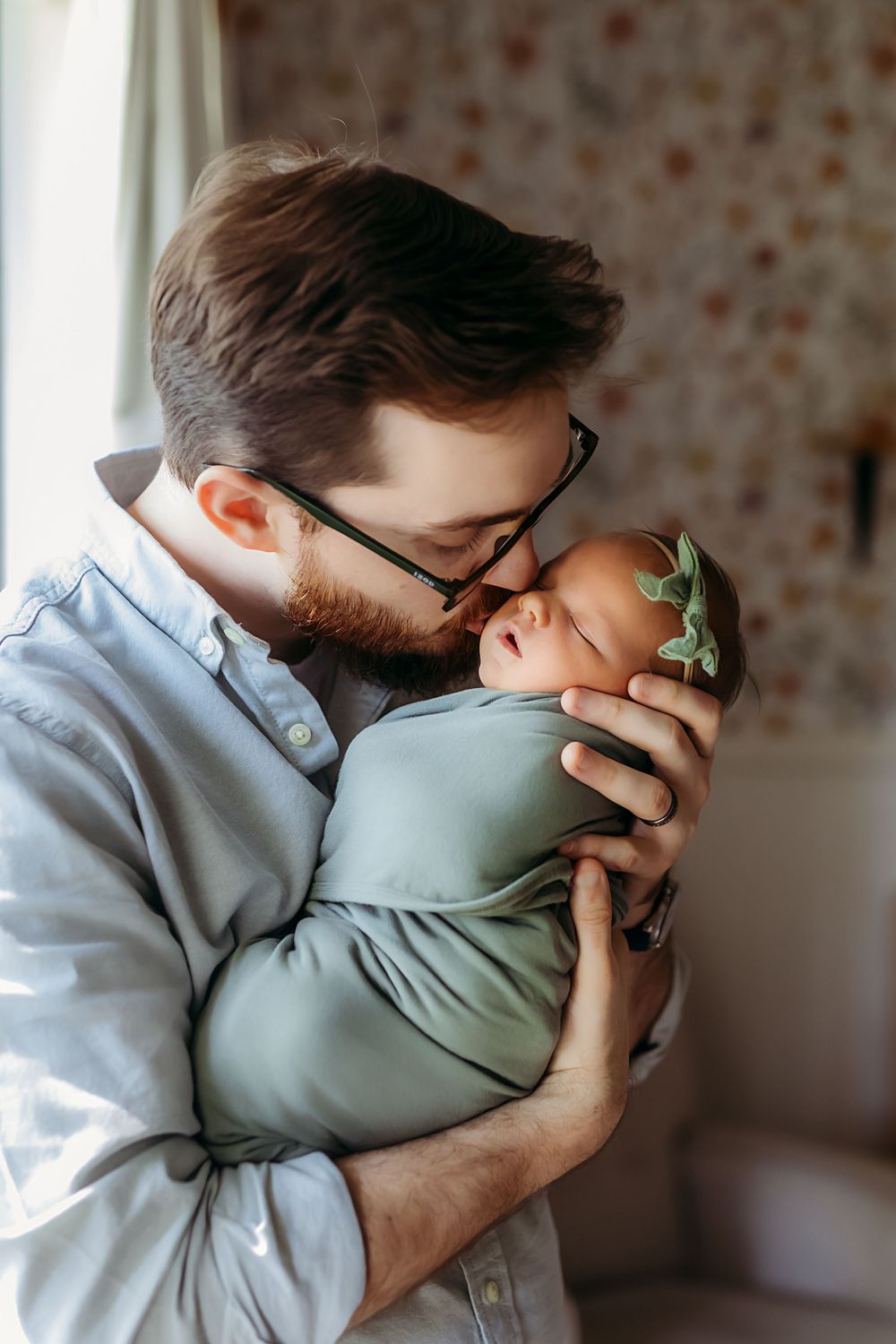 A tender moment captured as a bearded parent in glasses gently cuddles their swaddled baby in sage green.
