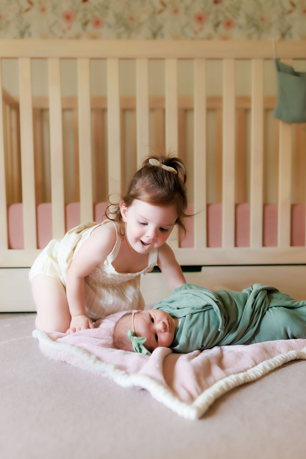 Two young siblings share a sweet moment together on a blanket beside a wooden crib.