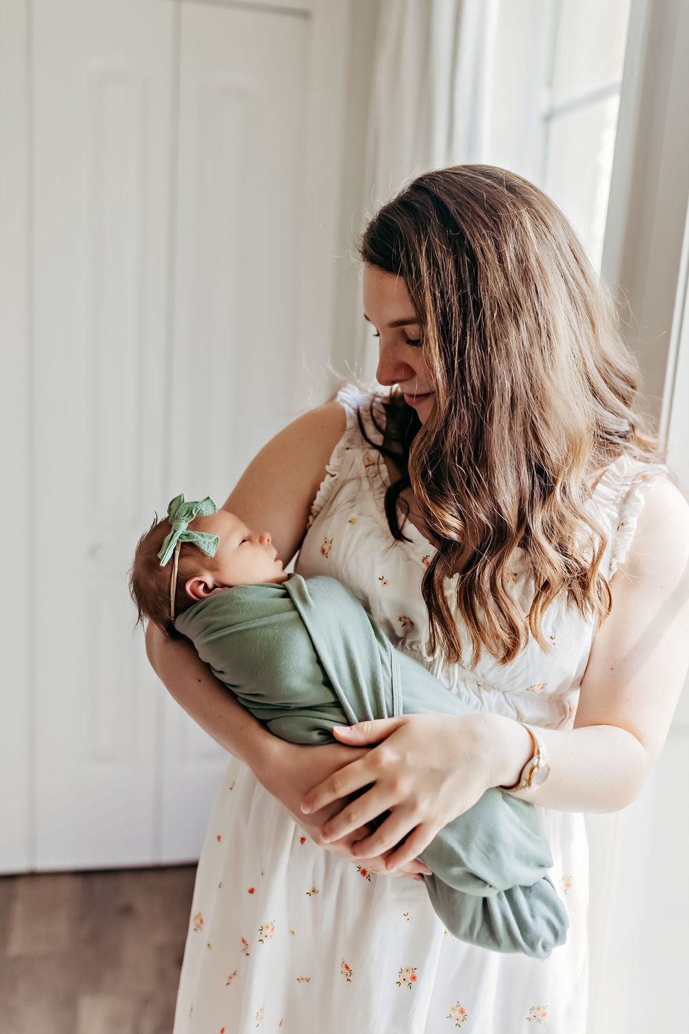 Mother cradles and kisses newborn baby wrapped in sage green swaddle against white dreamy backdrop.
