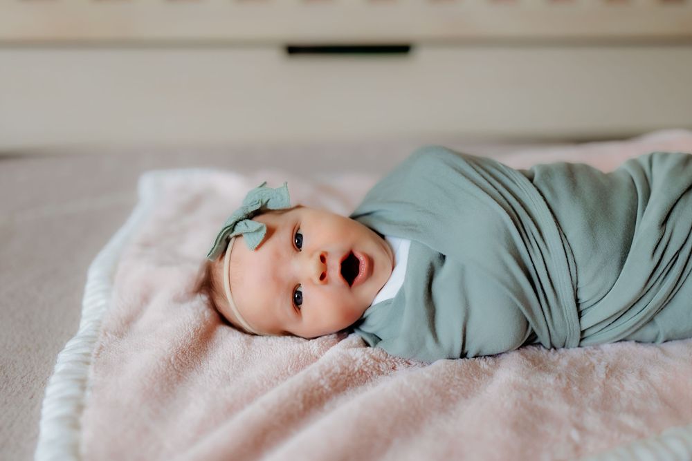 A smiling baby wrapped in a sage green swaddle blanket lies on a soft pink surface.