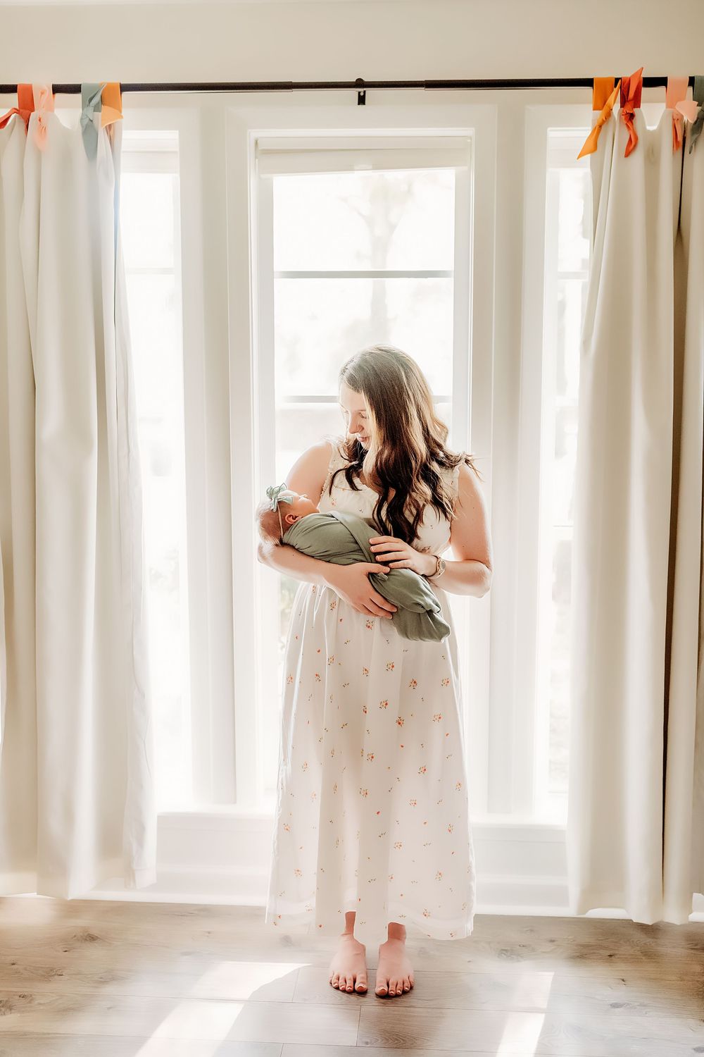 A peaceful nursery scene with sunlit windows, white curtains and parent holding infant in sage green swaddle.