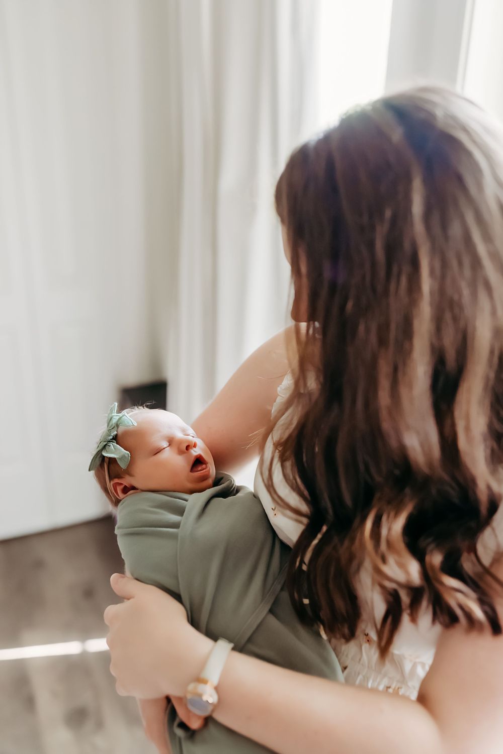 A newborn baby swaddled in olive green fabric yawns while being held in sunlit room.