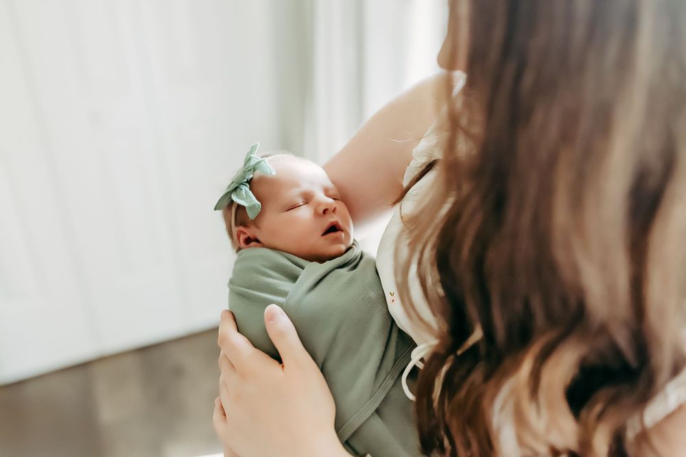 Mother lovingly embraces tiny newborn baby dressed in soft sage green in bright natural light setting.