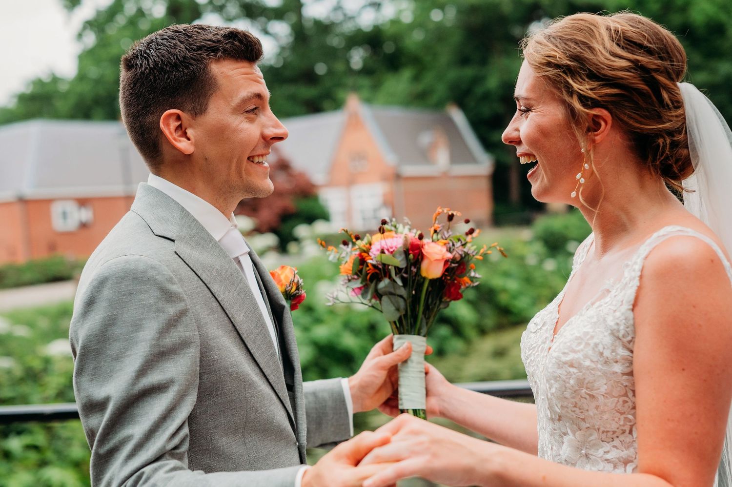A couple shares a joyful moment exchanging flowers on their wedding day in an outdoor garden setting.