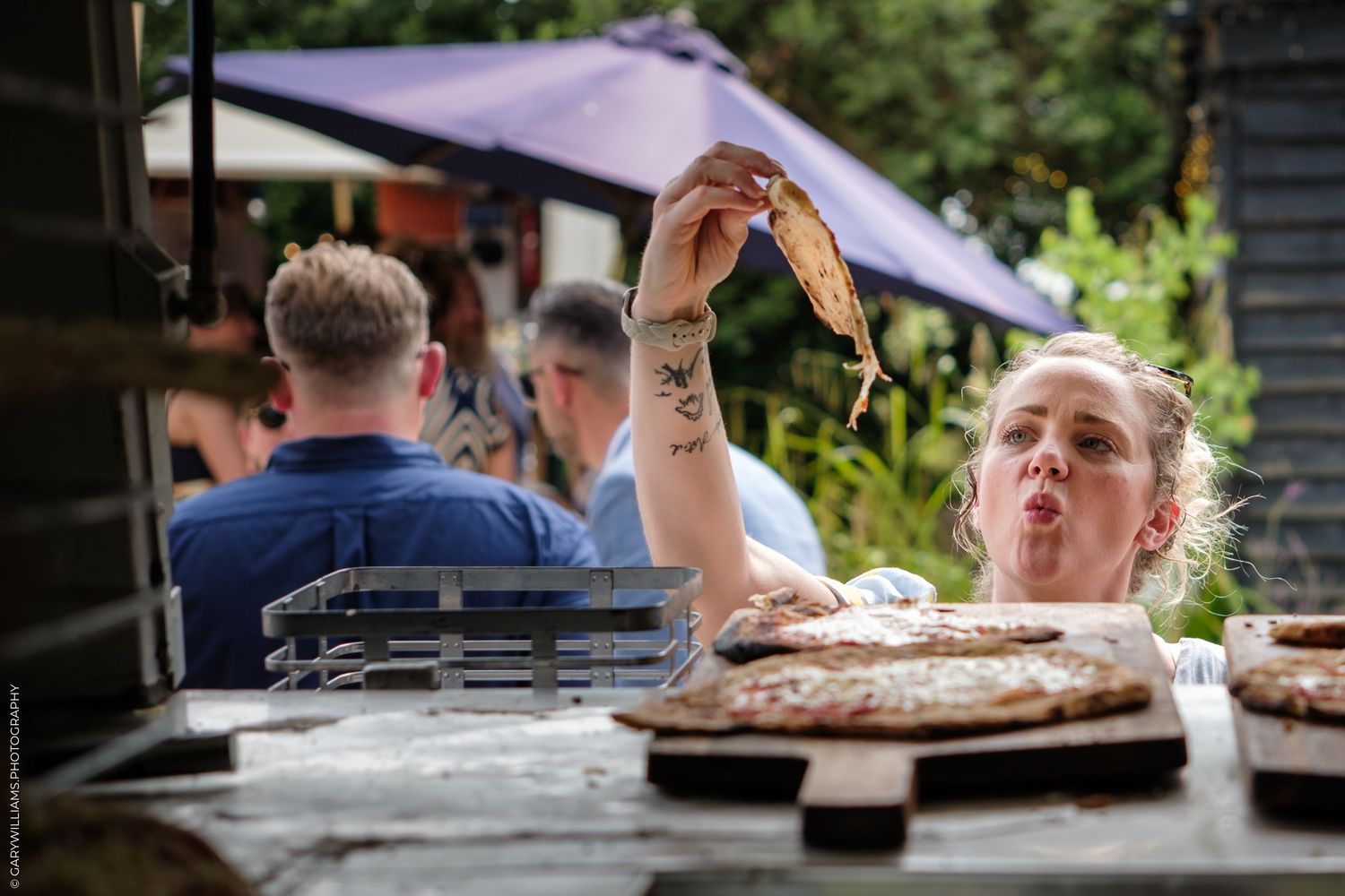An energetic chef stretches pizza dough in the air while preparing food outdoors at a restaurant patio.