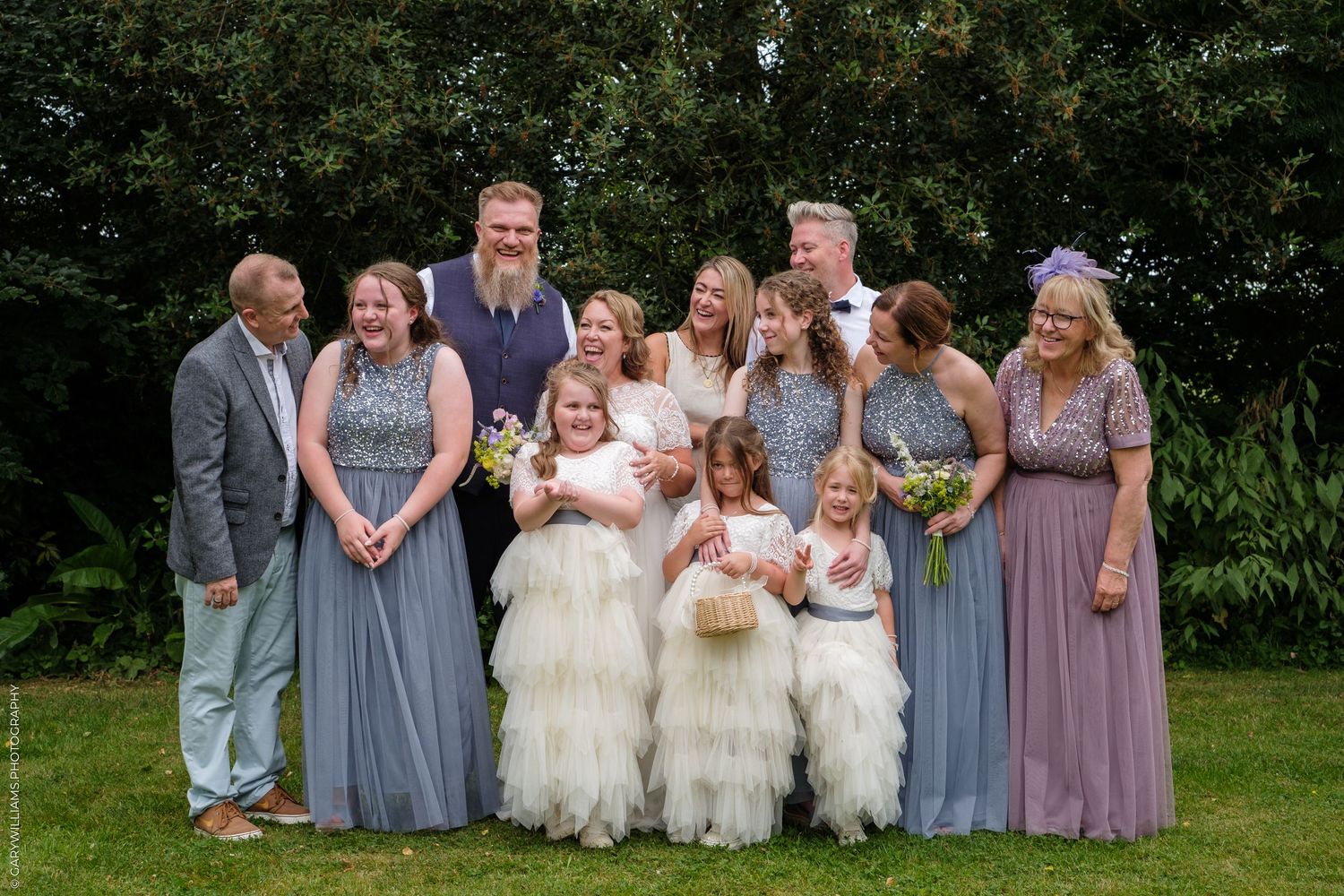 Wedding party members in blue-gray dresses and flower girls in white dresses pose together for outdoor group photos.