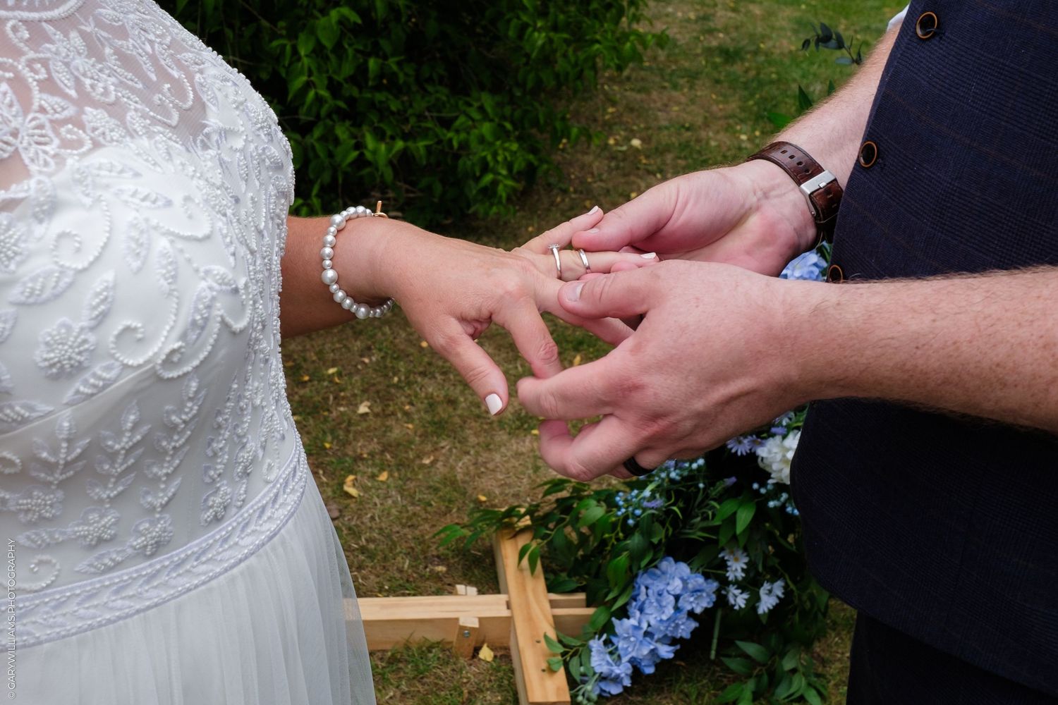 Close-up of hands exchanging wedding rings during outdoor ceremony with white lace dress and blue flowers visible.