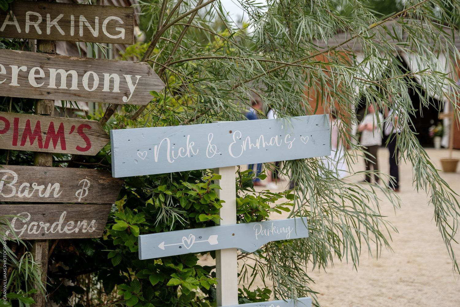 Rustic wooden directional signs for a wedding ceremony and venue among green foliage.